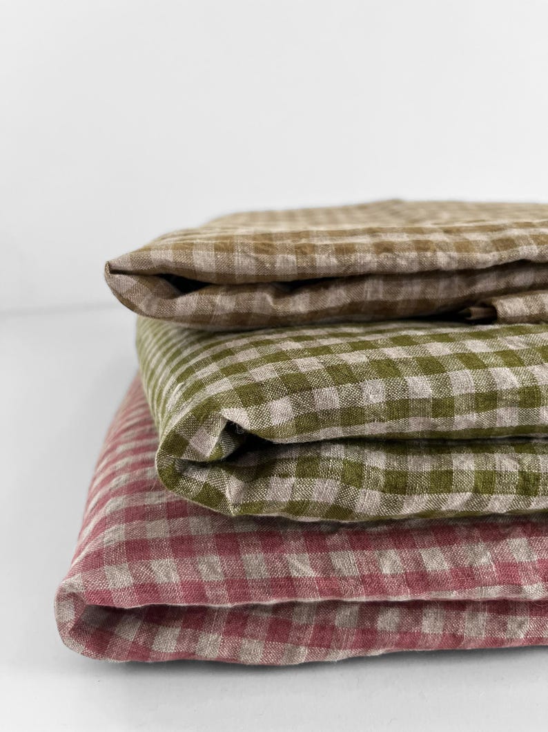 May include: A stack of three folded linen textiles in a gingham pattern. The top textile is a muted brown, the middle is olive green, and the bottom is a dusty rose color. The fabrics are neatly folded, showcasing the texture and pattern.