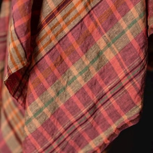 May include: Close-up of a fabric with a plaid pattern. The fabric features a mix of colors, including shades of pink, orange, green, and brown. The plaid design is made up of intersecting lines, creating a classic and timeless look. The fabric appears to be soft and lightweight.