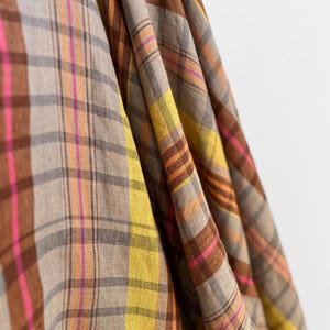 May include: Close-up of a fabric with a plaid pattern. The fabric features a mix of colors, including brown, tan, yellow, pink, and orange. The plaid design is made up of intersecting lines that create a grid-like pattern. The fabric is draped, showing the texture and folds.