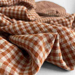 May include: Close-up of a folded fabric with a classic orange and white gingham pattern. The fabric appears to be linen or a similar natural fiber, with a soft, textured appearance. The image suggests a textile product, possibly for home decor or apparel.