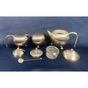 May include: A brushed stainless steel tea set consisting of a teapot with a lid, a creamer with a lid, a sugar bowl with a lid, a mesh tea infuser, and a small spoon. Each piece features a rounded body and stands on three small spherical feet.