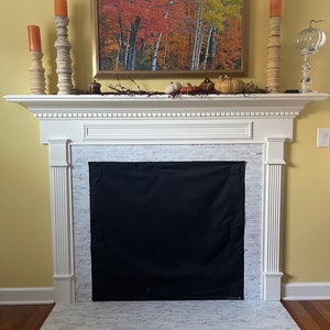 May include: A white fireplace mantel features a black fabric fireplace cover over the hearth. The mantel is decorated with three tall wooden candle holders with orange candles, a framed autumn forest painting, and small decorative pumpkins.