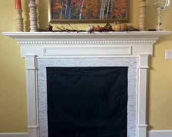 May include: A white fireplace mantel features a black fabric fireplace cover over the hearth. The mantel is decorated with three tall wooden candle holders with orange candles, a framed autumn forest painting, and small decorative pumpkins.