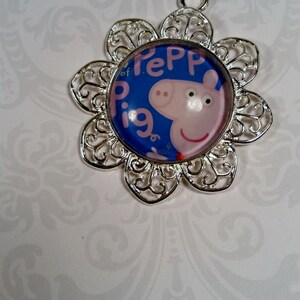 May include: A silver charm with a blue and pink Peppa Pig image in the center. The charm is shaped like a flower with intricate details.