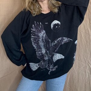 May include: Black long-sleeve sweatshirt featuring a large, detailed graphic of an eagle in flight. The eagle is depicted in shades of gray and white against a black background. The word "Alaska" is printed above the eagle. A moon graphic is also present.