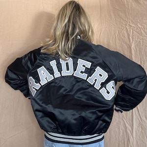 Puede incluir: Chaqueta de satén negra con la palabra "RAIDERS" arqueada en la espalda en letras blancas y grises. La chaqueta tiene un dobladillo y puños a rayas. Chaqueta de estilo clásico.