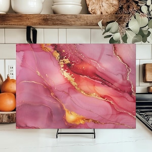 May include: A rectangular decorative glass chopping board with a pink and gold abstract design. The pink hues blend with gold accents, creating a marbled effect. The board is displayed on a black metal stand.
