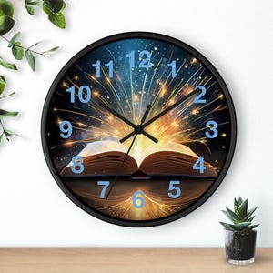 May include: A round black wall clock featuring a fantasy book design. The clock face has blue numerals and black hands. The open book is in the center, with a burst of light emanating from it. The clock is on a wooden surface.