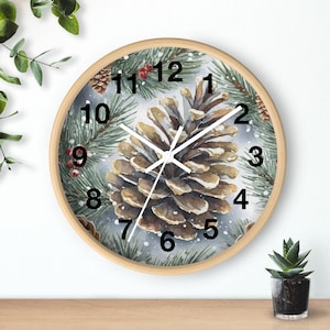May include: A round wall clock with a light wood frame. The clock face features a watercolor painting of a pine cone surrounded by pine needles and berries. Black numerals mark the hours.