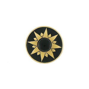 May include: A round, black and gold decorative knob with a starburst design. The knob features a central black circle surrounded by a gold star pattern on a black background. The knob is likely made of metal.