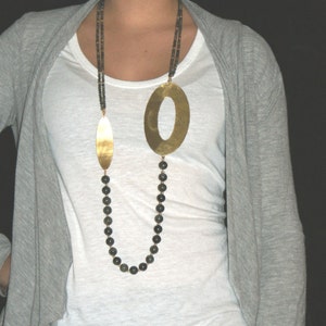 May include: A long necklace with a gold oval pendant and a strand of dark grey beads. The necklace is worn by a person with a grey cardigan and a white t-shirt.