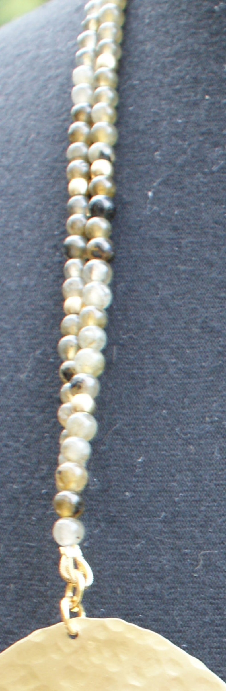 May include: A gold necklace with a large, round pendant. The necklace is made of multiple strands of small, round, white and grey beads.