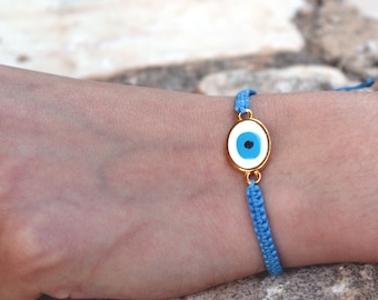 May include: A blue string bracelet with a gold evil eye charm. The charm is oval shaped with a white background and a blue eye with a black pupil.