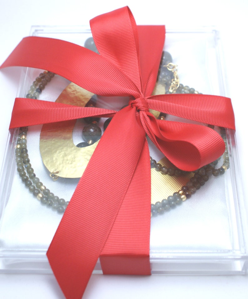 May include: A clear acrylic box with a red ribbon bow, containing a gold necklace with grey beads and a gold pendant.