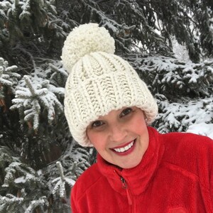 May include: A white knitted beanie with a large white pom pom. The beanie is worn by a person with a red fleece jacket. The person is standing in front of a snowy evergreen tree.