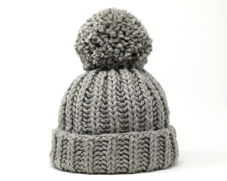 May include: A gray knitted beanie with a fluffy pom pom on top. The beanie has a ribbed texture.