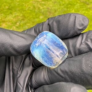 May include: A translucent, rounded gemstone with a blend of blue and white hues. The stone is held in a gloved hand, showcasing its polished surface and natural variations. The gem's color shifts from light blue to a milky white.