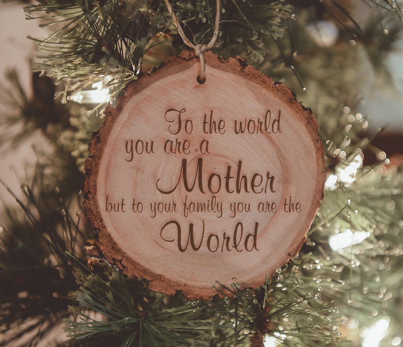 Puede incluir: Un adorno de madera con el texto "To the world you are a Mother but to your family you are the World" colgado de un &aacute;rbol de Navidad.