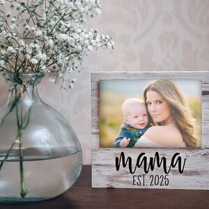 May include: A wooden picture frame with a photo of a mother and child. The frame is white with a distressed finish and the word "mama" and "EST. 2025" in black script. A clear glass vase with white flowers is to the left.
