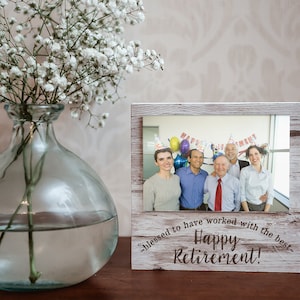 May include: A wooden picture frame with a photo of a group of people, with the text "Happy Retirement!" and "blessed to have worked with the best." A clear glass vase with white flowers is to the left.