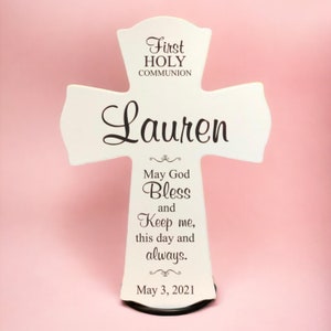 May include: White wooden cross with the words "First Holy Communion" and the name "Lauren" engraved on it. The cross also has the text "May God Bless and Keep me, this day and always." and the date "May 3, 2021".