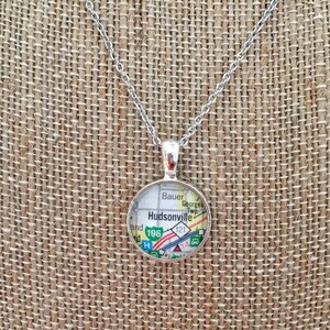 May include: Silver chain necklace with a round pendant featuring a map of Hudsonville, Michigan. The pendant includes the text "Hudsonville" and the highway numbers "196" and "121".