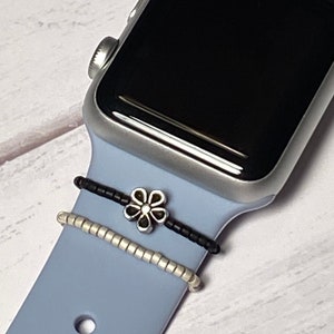 May include: A silver Apple Watch with a light blue silicone band. The band has two silver beaded bracelets and a silver flower charm.