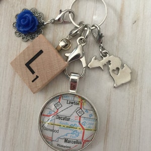 May include: A silver charm necklace with a map charm featuring the town of Marcellus, Michigan. The necklace also includes a wooden block with the letter "L", a blue flower charm, and a silver charm shaped like the state of Michigan.