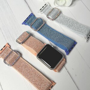 May include: Four Apple Watch bands in shades of silver, blue, and rose gold. Each band features a sparkly, textured fabric and a rectangular buckle adorned with iridescent rhinestones. The bands are displayed on a white wooden surface.