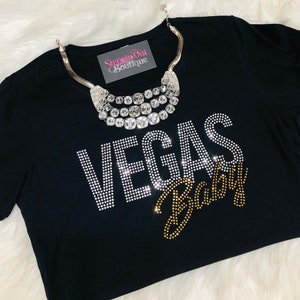 Pu&ograve; includere: Maglietta nera con decorazioni di strass che compongono la scritta "Vegas Baby" in oro e argento. La maglietta &egrave; appoggiata su una superficie bianca e soffice con una collana d'argento sopra.