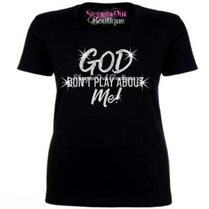 May include: Black t-shirt with the text "GOD DON'T PLAY ABOUT ME!" in a sparkling, rhinestone-like font. The shirt also features the text "Steppin Out Boutique" in a cursive font at the top.