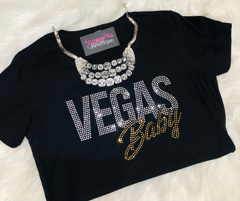 Pu&ograve; includere: Maglietta nera con decorazioni di strass che compongono la scritta "Vegas Baby" in oro e argento. La maglietta &egrave; appoggiata su una superficie bianca e soffice con una collana d'argento sopra.