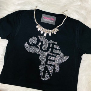 Op de afbeelding: Zwart T-shirt met een strass-ontwerp van het Afrikaanse continent met het woord "QUEEN" uitgespeld binnen de omtrek. Het shirt heeft een zilveren ketting met rechthoekige schakels.