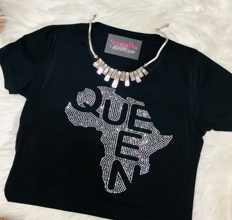 Op de afbeelding: Zwart T-shirt met een strass-ontwerp van het Afrikaanse continent met het woord "QUEEN" uitgespeld binnen de omtrek. Het shirt heeft een zilveren ketting met rechthoekige schakels.