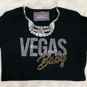 Pu&ograve; includere: Maglietta nera con strass che compongono la scritta "Vegas Baby" in un design oro e argento. La maglietta &egrave; indossata con una collana argentata a pi&ugrave; strati.
