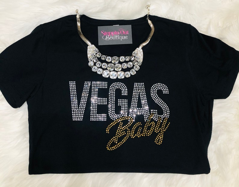 Pu&ograve; includere: Maglietta nera con strass che compongono la scritta "Vegas Baby" in un design oro e argento. La maglietta &egrave; indossata con una collana argentata a pi&ugrave; strati.