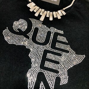 Op de afbeelding: Zwart T-shirt met een strass-ontwerp van het woord "QUE" en een zilveren ketting met een geometrisch ontwerp. Het shirt heeft de tekst "Steppin Out Boutique" op een wit label.