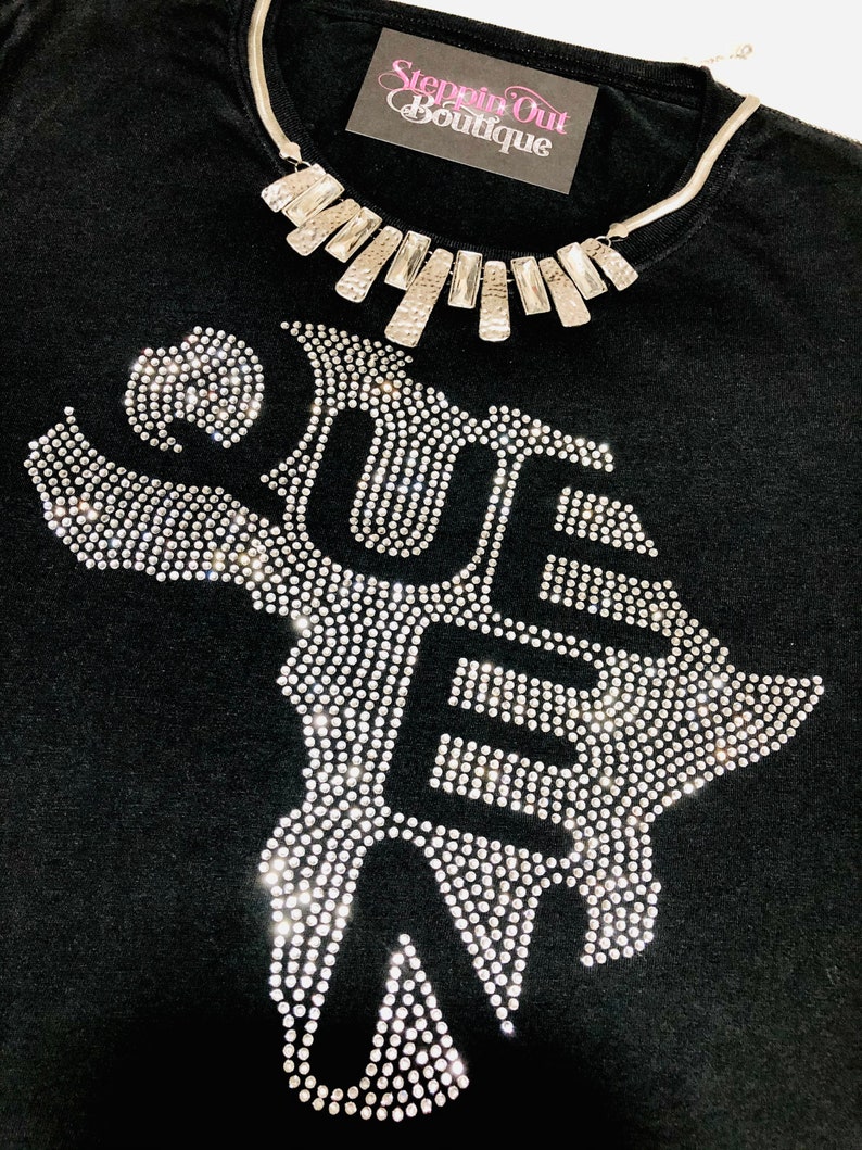 Op de afbeelding: Zwart T-shirt met een strass-ontwerp van het woord "QUE" en een zilveren ketting met een geometrisch ontwerp. Het shirt heeft de tekst "Steppin Out Boutique" op een wit label.
