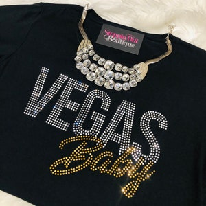 Pu&ograve; includere: Maglietta nera con decorazioni in strass che compongono la scritta "Vegas Baby" e una collana di strass argentata. La maglietta ha la scritta "Steppin Out Boutique" stampata su un'etichetta.