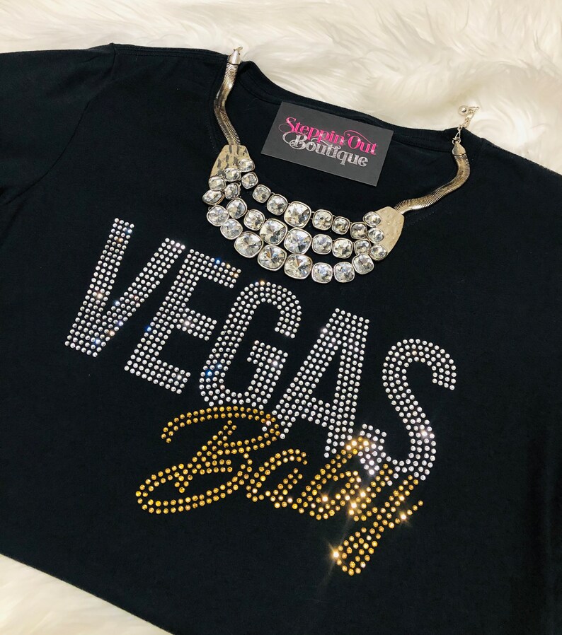 Pu&ograve; includere: Maglietta nera con decorazioni in strass che compongono la scritta "Vegas Baby" e una collana di strass argentata. La maglietta ha la scritta "Steppin Out Boutique" stampata su un'etichetta.