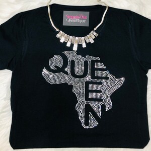 Op de afbeelding: Zwart T-shirt met een strass-ontwerp van het Afrikaanse continent en het woord "QUEEN" erop. Het shirt heeft een zilveren ketting met rechthoekige bedels.