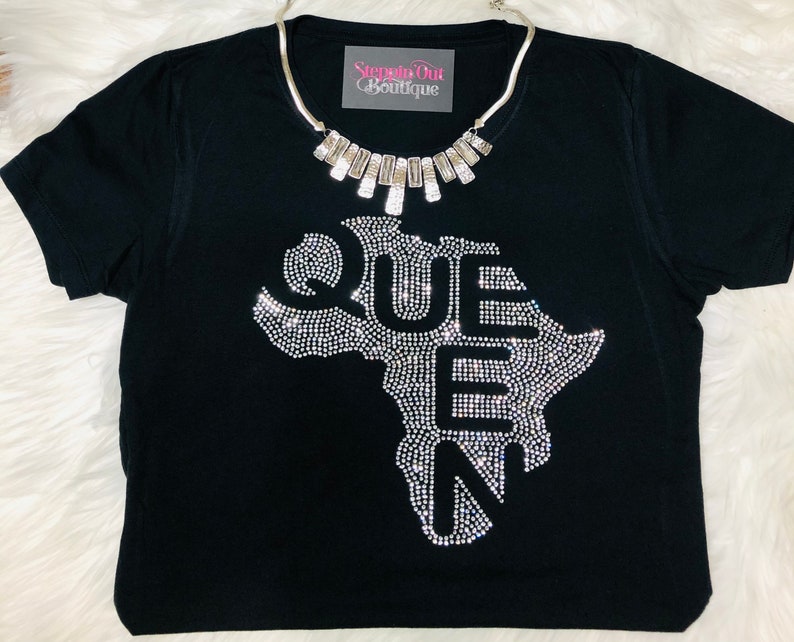 Op de afbeelding: Zwart T-shirt met een strass-ontwerp van het Afrikaanse continent en het woord "QUEEN" erop. Het shirt heeft een zilveren ketting met rechthoekige bedels.