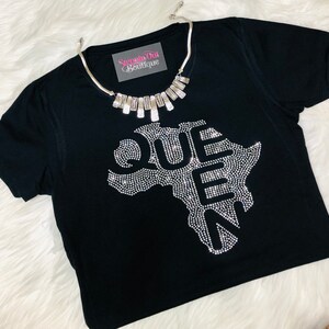 Op de afbeelding: Zwart T-shirt met een strass-ontwerp van het Afrikaanse continent met het woord "QUEEN" in strass. Het shirt heeft een zilveren ketting met een rechthoekige hanger met de tekst "Step Out Boutique".