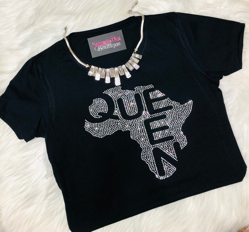 Op de afbeelding: Zwart T-shirt met een strass-ontwerp van het Afrikaanse continent met het woord "QUEEN" in strass. Het shirt heeft een zilveren ketting met een rechthoekige hanger met de tekst "Step Out Boutique".