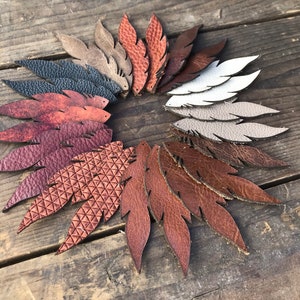 May include: A collection of leather feather shapes in various colors, arranged in a circular pattern. The feathers range in color from white to dark brown, with textures including smooth and embossed patterns. The arrangement is set against a rustic wooden surface.