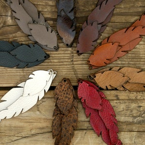 May include: A set of ten leather feathers in various colors, including white, black, brown, red, and orange. The feathers are arranged in a circle on a wooden surface.
