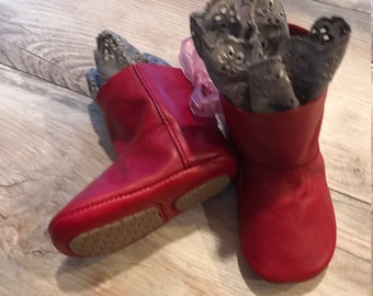 May include: A pair of bright red leather-like booties with a smooth finish and a rounded toe. Each bootie features a tall, wide cuff lined with a grey, laser-cut floral pattern fabric. The soles are tan with a textured, grid-like grip pattern.