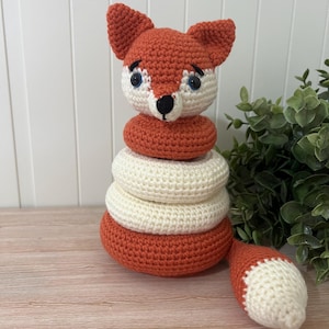 Puede incluir: Un juguete apilable de zorro hecho a mano. El zorro tiene una cabeza y orejas de color naranja rojizo, cara blanca y nariz negra. Los anillos apilables son blancos y naranja rojizo. La cola del zorro es blanca y naranja rojizo.