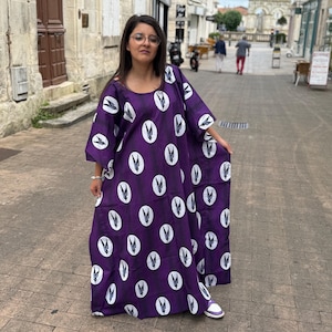 Peut inclure: Une longue robe violette fluide avec un motif ovale blanc. La robe a des manches courtes et une encolure arrondie. La femme porte des baskets blanches.