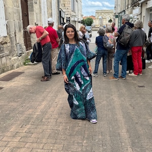 Peut inclure: Une femme porte une longue robe à motifs bleus et verts, debout dans une rue pavée. La robe a une coupe ample et des manches larges. D'autres personnes et des bâtiments sont visibles en arrière-plan.